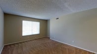 This single-floor residential space offers a straightforward and functional layout designed for comfortable living. The main living area is finished with wall-to-wall carpeting in a warm tan tone, providing a cozy underfoot feel throughout the space. Neutral beige walls create a clean, versatile backdrop that accommodates a variety of interior styles and furnishings. The room features a large window dressed with horizontal blinds, allowing natural light to fill the space while maintaining privacy. A textured popcorn ceiling adds a classic character to the room's overhead finish. The central HVAC vent positioned along the upper wall ensures efficient climate control throughout the unit. White baseboards provide a crisp contrast against the neutral walls, contributing to a tidy and well-defined aesthetic. Electrical outlets are conveniently distributed along the walls to support everyday functionality. The overall ambiance of the space is clean, understated, and move-in ready, offering a neutral canvas well-suited for personalization. The single-floor configuration provides ease of movement and accessibility throughout the home.