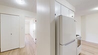 This thoughtfully laid-out 1-bedroom, 1-bathroom unit offers a clean, modern aesthetic across a single floor. Light-toned hardwood flooring flows throughout the space, creating a cohesive and airy feel. The open living area is generously sized, featuring a baseboard heating unit along the wall and a neutral white palette that enhances the sense of openness. A hallway connects the main living area to the private spaces, including the bedroom and bathroom.

The kitchen is efficiently designed, featuring white cabinetry with upper storage extending to the ceiling, a full-size refrigerator, and a compact stovetop, making practical use of the available space.

The bedroom offers a comfortable footprint with a large window fitted with horizontal blinds that invite natural light while providing privacy. Outdoor views of surrounding greenery can be glimpsed through the window. A built-in closet with folding doors provides ample storage.

The bathroom continues the clean, functional theme with a granite-topped vanity, an undermount sink, and a large mirror. Adjacent to the bathroom, an additional closet with shelving and a hanging rod offers further storage convenience.

Throughout, the textured ceilings and crisp white walls maintain a consistent, understated finish that gives the space a fresh, move-in-ready character.