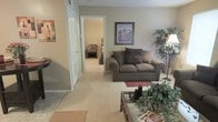 This single-floor space features an open and functional layout centered around a carpeted living area finished in warm, neutral tones. The living room is furnished with a plush, earth-toned loveseat accented with decorative pillows, complemented by a glass-top coffee table and a red accent rug that adds a subtle pop of color. Framed wall art depicting a scenic landscape enhances the warm aesthetic, while a large decorative plant adds a natural, organic element to the corner of the room. The flooring transitions from carpet in the main living area to tile near the entryway, providing practical durability at the threshold. An open doorway leads directly into a bedroom, which is visible from the living space and continues the neutral color palette with matching carpet. Interior doors with white trim provide access to additional spaces, suggesting a closet or hallway beyond. The overall ambiance is understated and comfortable, with a cohesive color scheme of tans and greens throughout, creating a welcoming and livable environment.