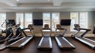 This fitness center is designed to accommodate a comprehensive range of workout styles within a single, open-plan layout. The space is finished with soft blue-gray walls, white crown molding, tray ceilings, and rubber flooring throughout, creating an environment that balances functionality with polished aesthetics. Ceiling fans are positioned throughout, supporting airflow and comfort during workouts.

The cardio area lines one wall with multiple treadmills positioned in front of large windows, each paired with wall-mounted flat-screen televisions. Natural light streams in from multiple windows across the facility, offering views of an outdoor pool area and surrounding landscaping. A dedicated section houses stationary bikes, a recumbent bike, and a rowing machine, also served by windows and additional wall-mounted screens.

The strength and functional training zone includes a multi-station cable machine, a weight bench, dumbbell racks, medicine balls, exercise balls, a heavy bag, and a functional training rig. An adjacent corridor leads to additional amenities. The overall ambiance is bright, clean, and inviting, reflecting a thoughtfully designed community fitness facility.