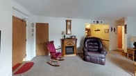 This single-floor, two-bedroom home offers a comfortable and inviting layout with a warm, homey character throughout. The spacious living room serves as the heart of the home, featuring a wood-trimmed electric fireplace with a decorative mantel, carpeted flooring, and an open passage to an adjacent sitting or family area. The living spaces are furnished with plush seating and accented with classic country-style décor, creating a cozy and relaxed atmosphere.

The galley-style kitchen is efficiently laid out with dark wood cabinetry, white appliances including a range and refrigerator, a double sink, and light tile flooring. Ample upper cabinet storage and a textured ceiling complete the functional design.

Both bedrooms are carpeted and feature warm tan-toned walls, natural light from curtained windows, and generous space for furnishings. One bedroom offers a traditional wood-framed bed with a classic dresser, while the second bedroom features a lighter, painted furniture set in a soft, relaxed style.

The full bathroom includes a tiled tub-shower combination, a vanity with storage, tile flooring, and decorative border detailing near the ceiling, offering a clean and well-appointed space.

Overall, this home presents a welcoming, traditional aesthetic with practical amenities throughout.