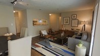 This single-story space offers an open and functional layout centered around a comfortable living area. The living room features hardwood-style flooring and is furnished with a mix of mid-century modern and contemporary pieces, including leather accent chairs, a dark wood coffee table, and a plush sofa. A bold geometric area rug in blue, white, and earth tones anchors the seating arrangement, adding a vibrant focal point to the room. A low-profile wooden credenza along the back wall provides additional storage and display space, complemented by a curated gallery of framed artwork above it.

A pass-through window connects the living area to the kitchen, enhancing the open flow between spaces. The kitchen, visible through the pass-through, features light cabinetry, a sink positioned beneath a window, and counter seating that allows for casual dining or conversation. The neutral, warm-toned walls throughout create a cohesive and inviting ambiance. The textured ceiling adds a subtle architectural detail, while interior lighting ensures the space remains well-lit. Overall, the layout balances practicality with a stylish, lived-in aesthetic.