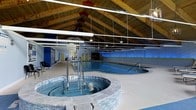 Located at 1600 Bonwood Road in Wilmington, Delaware, this single-floor indoor aquatic facility offers a well-appointed environment designed for both fitness and relaxation. The centerpiece of the space is a full-length lap pool with clearly marked lane lines, ideal for structured swim training or recreational use. Adjacent to the pool sits a generously sized circular hot tub spa, elegantly bordered by a natural stone surround with stainless steel handrails for safe entry and exit.

The architecture of the facility is a standout feature. A soaring vaulted ceiling with exposed natural wood beams and tongue-and-groove wood planking creates a warm, lodge-like ambiance that contrasts beautifully with the cool blue tones of the water below. Pendant-style industrial lighting and overhead tube lights ensure consistent, bright illumination throughout the space.

The walls are finished in a bold blue and white color scheme, accented by blue LED lighting that reflects dramatically off the pool water, enhancing the atmosphere. A separate anteroom or utility corridor is visible at one end, providing access to support facilities. The overall design balances functional aquatic infrastructure with an inviting, resort-like aesthetic, making it a versatile space suited for therapeutic use, fitness programming, or leisure swimming.