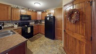 This single-floor home exudes a warm, rustic charm throughout its well-appointed living spaces. The main living area is generously sized, featuring plush carpeting, a corner stone fireplace with a wooden mantle, and large sliding glass doors that allow abundant natural light and provide views of the outdoor greenery. The space is styled with farmhouse-inspired décor, including decorative wall signage, vintage accents, and an Edison bulb pendant light that adds a distinctive touch.

A second sitting area connects seamlessly to the main living room, offering built-in shelving, soft carpet flooring, and additional seating — ideal for relaxation or entertaining. The kitchen is well-equipped with rich knotted wood cabinetry, stone-tile flooring, black appliances including a range and full-size refrigerator, and a rustic wooden entry door adorned with a decorative wreath, reinforcing the home's country aesthetic.

The bathroom features warm wood cabinetry with ample storage, a vanity with a large framed mirror, tile mosaic flooring, and a shower-tub combination. Throughout the home, the warm beige and tan color palette, textured ceilings, and carefully curated décor create a cohesive, inviting atmosphere that blends comfort with rustic, farmhouse-style character.