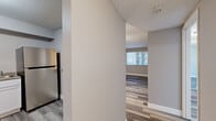 This single-floor, 2-bedroom, 2-bathroom home offers a clean and modern interior with consistent light wood-look flooring running throughout every room. The living room is a generous, open space featuring a ceiling fan with a light fixture, neutral gray walls, and a window that allows natural light to fill the area. A pass-through counter connects the living area to the kitchen, which is equipped with white shaker-style cabinetry, granite-look countertops, a double stainless steel sink, and an electric range — providing both style and practicality. The primary bedroom is a spacious retreat with a window fitted with blinds, offering ample room for furnishings. The second bedroom mirrors the same neutral tones and wood-look flooring, also featuring a window with blinds. Both bathrooms are functional and clean, with one featuring a single-sink vanity with chrome fixtures, a medicine cabinet mirror, and a full-sized tub/shower combo with white surround panels. A hallway connects the bedrooms and bathrooms, maintaining a logical and efficient floor plan. The overall design is cohesive and contemporary, with neutral warm-gray walls and bright white trim creating a welcoming, move-in-ready atmosphere throughout.