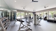 This fitness center offers a comprehensive, single-floor layout designed to support a full range of workout activities. The main exercise floor is generously proportioned, featuring durable rubberized flooring and an impressive array of strength and cardio equipment, including cable machines, ellipticals, stair climbers, rowing machines, and stationary bikes. Full-length mirrors line the walls, enhancing the sense of openness, while multiple ceiling fans and recessed lighting keep the environment comfortable and well-lit. Large floor-to-ceiling windows and glass doors run along the perimeter, flooding the space with natural light and offering views of an outdoor pool area and surrounding landscaped grounds. A dedicated free weights section houses a full rack of dumbbells alongside a weight bench, providing a focused space for strength training. Adjacent to the main floor, a separate room contains a stand-up tanning bed unit with light wood-look flooring, offering a private wellness amenity. A hallway area near the entrance features additional private rooms accessible via paneled gray doors, along with a seating area with a modern accent chair. The overall ambiance is clean, modern, and functional, making this facility a well-rounded destination for fitness and wellness.