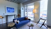 This single-floor space showcases a compelling blend of industrial and modern design elements. The living area draws immediate attention with its exposed concrete beam ceiling, which adds a distinctive architectural character and an urban loft-like quality to the interior. Large floor-to-ceiling windows flood the space with natural light and offer open views of the surrounding cityscape, creating a strong visual connection to the outside environment.

The living area is well-appointed with a bold blue velvet sofa accented by decorative throw pillows, anchored on a patterned area rug. Natural wood slice coffee tables provide an organic contrast to the industrial ceiling above. A sleek metal shelving unit displays curated décor, including a wall-mounted wine rack and framed artwork, reinforcing the space's contemporary sensibility. A tripod floor lamp adds both functional lighting and a sculptural element to the room.

The overall ambiance is modern and curated, balancing raw industrial finishes with carefully selected furnishings. The interplay of textures—concrete, velvet, wood, and metal—creates a layered, sophisticated atmosphere that feels both stylish and livable.