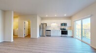 This single-floor unit showcases a clean, contemporary design with light-toned luxury vinyl plank flooring running consistently throughout the space. The open-concept layout connects the kitchen and living area seamlessly, creating a bright and functional environment. The kitchen is well-appointed with white shaker-style cabinetry, a full suite of stainless steel appliances including a refrigerator, dishwasher, electric range, and over-the-range microwave, along with a single-basin sink and recessed lighting overhead.

The living area is generously sized, benefiting from natural light and direct access to a private balcony through a sliding glass door — the balcony also features an adjacent window, enhancing the sense of openness. A dedicated bedroom offers ample space, with its own sliding glass door leading to the balcony and an additional window framing outdoor views of the surrounding community.

The full bathroom features a white vanity with black hardware accents, a tub-shower combination, and matching wood-look flooring. A hallway connects the main entry — fitted with a solid panel door — to the bedroom and bathroom, with additional storage visible via a walk-in closet area. The overall ambiance is bright, neutral, and move-in ready.
