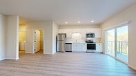 This single-floor unit showcases a clean, contemporary design with light-toned luxury vinyl plank flooring running consistently throughout the space. The open-concept layout connects the kitchen and living area seamlessly, creating a bright and functional environment. The kitchen is well-appointed with white shaker-style cabinetry, a full suite of stainless steel appliances including a refrigerator, dishwasher, electric range, and over-the-range microwave, along with a single-basin sink and recessed lighting overhead.

The living area is generously sized, benefiting from natural light and direct access to a private balcony through a sliding glass door — the balcony also features an adjacent window, enhancing the sense of openness. A dedicated bedroom offers ample space, with its own sliding glass door leading to the balcony and an additional window framing outdoor views of the surrounding community.

The full bathroom features a white vanity with black hardware accents, a tub-shower combination, and matching wood-look flooring. A hallway connects the main entry — fitted with a solid panel door — to the bedroom and bathroom, with additional storage visible via a walk-in closet area. The overall ambiance is bright, neutral, and move-in ready.