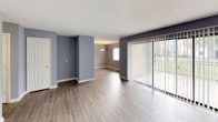 Aura is a well-appointed single-floor residence designed with a consistent and calming aesthetic throughout. The interior is unified by soft blue-grey walls and crisp white trim, creating a clean, modern atmosphere from room to room.

The open living area welcomes you with light wood-style laminate flooring that extends seamlessly into the adjoining dining space. A sliding glass door floods the area with natural light and provides outdoor access, while a pendant light fixture anchors the dining zone. The layout offers generous open space, ideal for comfortable everyday living.

The galley kitchen is efficiently designed with white cabinetry lining both walls, light laminate countertops, a double stainless steel sink, and black appliances including a refrigerator and range with overhead ventilation. A window allows natural light into the cooking space, adding to its functional appeal.

The bedrooms are carpeted for comfort and feature the same blue-grey color palette. One bedroom includes two closets — one walk-in and one standard — offering ample storage. Additional bedrooms provide windows for natural light and convenient access to shared spaces.

Two full bathrooms serve the residence, each with a tub-shower combination, vanity with mirror, and neutral finishes. The bathrooms are clean and practical in design, complementing the overall tone of the home.