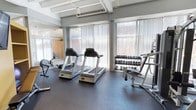 This fitness center is a purpose-built exercise space designed to support a comprehensive workout routine. The room is bright and airy, thanks to large floor-to-ceiling windows fitted with vertical blinds that allow generous natural light to filter in while offering privacy. The windows frame views of the surrounding brick exterior, adding a grounded, urban character to the space. Structurally, the room features exposed ceiling beams and visible ductwork, lending it an industrial aesthetic that complements its functional purpose. Sturdy columns are positioned throughout, reinforcing the open layout. The flooring is a dark, rubberized surface well-suited for exercise, providing durability and traction. Equipment includes multiple Matrix-brand treadmills positioned prominently in the center of the room, a rowing machine, and a multi-use weight bench. A well-stocked dumbbell rack lines the wall near the windows, offering a wide range of free weights for strength training. The overall ambiance is clean, practical, and well-maintained, making it a functional and inviting space for both cardio and strength-based exercise.