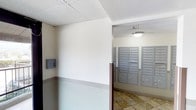 This single-floor common area serves as a functional shared space centered around a well-organized mail room. The space features a series of built-in, multi-unit metal mailboxes mounted along the walls, providing individual mail access for numerous occupants. The mailboxes are finished in a sleek gray tone, offering a clean and orderly appearance. The flooring is composed of stone-patterned tiles in warm, earthy tones, lending a grounded and durable quality to the space. Walls are painted in a soft neutral tone with a contrasting darker wainscoting border along the lower portion, adding a subtle architectural detail. The ceilings feature a textured tile finish, and the area is illuminated by a flush-mounted ceiling light that provides consistent, even lighting throughout. A wall-mounted intercom or access control panel is visible, indicating a secure, managed environment. The overall ambiance is practical and well-maintained, designed to efficiently serve the day-to-day needs of building residents with a straightforward, no-frills aesthetic that prioritizes functionality and ease of use.