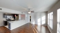 This single-floor residence offers an open-concept layout that seamlessly connects the living area to the kitchen, creating a spacious and airy environment. Rich, dark hardwood flooring runs throughout the main living space, adding warmth and a polished aesthetic to the interior.

The living area is generously proportioned, bathed in natural light streaming through blinded windows that cast a warm, patterned glow across the floors. A ceiling fan with an integrated light fixture is centrally mounted overhead, providing both comfort and ambient lighting. The neutral white walls complement the clean, contemporary feel of the space.

The kitchen is partially open to the living area via a pass-through counter with a light-colored stone or marble-style surface and a sink, making it functional and social. Pendant lighting in a bold red hue adds a striking design accent above the counter. The kitchen is equipped with stainless steel appliances, including a refrigerator, and features dark cabinetry that contrasts well with the lighter countertops.

A hallway leading off the main living space provides access to additional rooms, suggesting a well-structured and practical floor plan. An HVAC panel is neatly integrated into the wall near the hallway entry. Overall, the space delivers a modern, clean, and inviting atmosphere suited for comfortable everyday living.