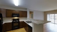This single-floor space is designed with practicality and comfort in mind, centered around an open-concept kitchen that serves as the heart of the layout. The kitchen is outfitted with rich, dark wood cabinetry offering generous storage, paired with light granite-style countertops that provide ample prep space. Black appliances, including an electric range, built-in microwave, and dishwasher, lend a cohesive and modern aesthetic to the cooking area. A peninsula with an integrated sink extends the counter space and creates a natural transition between the kitchen and the adjoining living area, making it ideal for both meal preparation and casual dining. The light hardwood flooring throughout adds warmth and continuity to the space, while the neutral, light-toned walls keep the interior feeling bright and open. Overhead lighting ensures the kitchen is well-lit, enhancing visibility across the workspace. A pass-through opening connects the kitchen to an adjacent room, further reinforcing the open and airy feel of the overall layout. The single-floor design ensures easy accessibility and a straightforward, well-connected flow throughout the entire space.