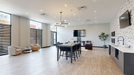 Located at 1179 E Main St in Columbus, Ohio, this contemporary community lounge offers a spacious, open-plan layout spread across a single floor. Light-toned hardwood flooring runs throughout, complementing the neutral white and gray walls to create a bright, welcoming atmosphere. Floor-to-ceiling windows with horizontal slatted screens line the exterior walls, allowing abundant natural light to filter in while adding a striking architectural element to the interior.

The space is thoughtfully organized into distinct functional zones. A central high-top dining table with dark woven chairs serves as a gathering and collaborative hub, anchored beneath a sculptural multi-globe chandelier that adds an artistic focal point overhead. Recessed lighting supplements the natural light throughout. A dedicated lounge area features a neutral sofa, dark accent chairs, a round side table, and a large wall-mounted flat-screen TV, creating a relaxed entertainment setting.

Additional seating vignettes with curved cream armchairs and minimalist side tables are positioned near the windows, ideal for casual conversation or individual use. Decorative wall art, including a set of sculptural face masks, adds cultural character to the space. A glass-partitioned entryway with black steel framing and a textured accent wall provides a polished, modern arrival experience. Overall, the space exudes a clean, sophisticated aesthetic suited for communal use.