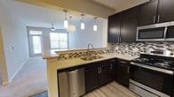 This single-floor residence offers a thoughtfully designed open-concept layout that promotes a sense of spaciousness and connectivity throughout. The kitchen serves as a central highlight, featuring rich dark cabinetry paired with light granite countertops that provide a striking contrast. A mosaic tile backsplash in neutral tones adds texture and visual interest to the cooking space. Stainless steel appliances, including a dishwasher and range, lend a sleek, contemporary feel, while a double-basin stainless steel sink with a gooseneck faucet anchors the workspace. A peninsula countertop with pendant lighting above creates a natural transition between the kitchen and the adjoining living area, offering both a casual dining or prep surface and an inviting gathering point. The pendant lights, varying in style yet cohesive in finish, provide warm focused illumination over the counter. The living area beyond benefits from abundant natural light streaming through large windows, complemented by a ceiling fan that enhances comfort and airflow. Light wood-toned flooring flows throughout, tying the spaces together with a clean, modern aesthetic. The overall ambiance is contemporary and welcoming, blending practical design with stylish finishes.
