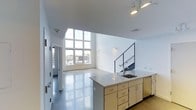 This contemporary two-story space showcases a clean, modern aesthetic defined by crisp white walls, polished concrete floors, and an abundance of natural light. The main level opens into a generous living area anchored by a dramatic floor-to-ceiling window grid that floods the interior with daylight and provides sweeping views of the surrounding urban landscape. The soaring ceiling height amplifies the sense of openness and volume throughout the space.

The open-plan kitchen features a central island with a granite countertop, an integrated sink, and ample cabinetry with light wood finishes and brushed metal hardware. Track lighting overhead provides focused illumination across the work surfaces. The kitchen flows seamlessly into the living area, making it well-suited for both everyday living and entertaining.

A modern staircase with a sleek metal railing connects the main level to the upper floor, adding an architectural focal point while maintaining a sense of visual openness. The polished concrete flooring throughout reflects light beautifully and contributes to the industrial-modern character of the space.

The layout also includes a corridor leading to additional rooms, suggesting versatile functionality across both floors. Overall, this space combines contemporary design, thoughtful materials, and an airy atmosphere to create a striking and highly livable environment.
