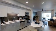 This single-floor space showcases a thoughtfully designed open-concept layout that seamlessly connects the kitchen, dining, and living areas. The kitchen is outfitted with stainless steel appliances, including an electric range, built-in microwave, and dishwasher. Crisp white upper cabinetry contrasts with gray lower cabinets, while a dark tile backsplash adds texture and visual depth. Light-toned countertops and a single-basin undermount sink complete the functional workspace. Recessed lighting illuminates the kitchen and flows throughout the unit, complemented by a ceiling fan in the living area. A compact round dining table with white chairs sits adjacent to the kitchen, anchored by a decorative plant arrangement. The living area features a darkened accent wall, creating a cozy focal point, and opens toward floor-to-ceiling windows that flood the space with natural light and offer views of the surrounding cityscape. Light wood-style flooring runs throughout, unifying the open layout and contributing to the bright, contemporary ambiance of the space.