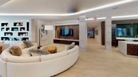 Located at 101 W Brorein St in Tampa, Florida, this single-floor amenity space delivers a refined, contemporary living experience defined by high-end finishes and thoughtful design. Light hardwood flooring runs throughout, unifying the open-concept layout and complementing the bright, neutral palette.

The main lounge area is anchored by a striking curved white sectional sofa positioned around a herringbone-patterned wood feature wall with a mounted flat-screen TV and recessed cove lighting that adds a soft, ambient glow to the curved ceiling above. A rustic live-edge wood stump serves as a natural accent piece, balancing the modern aesthetic.

An adjacent social lounge showcases a bold built-in shelving wall with dark niches displaying curated décor, while floor-to-ceiling windows flood the space with natural light and offer lush outdoor views of tropical greenery. A dramatic dining nook features a sculptural pendant light, a walnut dining table, and cream upholstered chairs, creating an intimate yet elegant setting.

The gourmet kitchen is outfitted with a sleek white waterfall island, bar seating, integrated appliances, and a textured mosaic tile accent wall that adds warmth and depth. Artistic touches—including a tall sculptural figure and a vibrant floral canvas—elevate the space's gallery-like ambiance throughout.