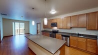 This single-floor residence offers a thoughtfully designed open-concept layout that seamlessly connects the kitchen and living areas. The kitchen is a standout feature, showcasing warm honey-toned wood cabinetry extending to the ceiling for ample storage, dark gray countertops, and a full suite of stainless steel appliances including a refrigerator, electric range, built-in microwave, and dishwasher. A central island with a matching countertop provides additional workspace and casual dining potential, while pendant lighting adds a refined touch overhead. Light hardwood flooring flows throughout the kitchen and living areas, enhancing the bright, airy atmosphere. The living room is generously proportioned with access to an outdoor balcony, allowing natural light to fill the space. The bedrooms feature plush neutral carpeting, providing a comfortable contrast to the hard flooring elsewhere, and include windows with blinds. Both bathrooms are finished consistently with warm wood vanity cabinetry, white countertops, large mirrors, and hardwood-style flooring. Wall-mounted medicine cabinets and grab bars provide functional detail. Overall, the space presents a clean, practical design with cohesive warm-toned finishes throughout.