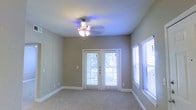 This single-floor space offers a clean, comfortable layout designed with both function and style in mind. The interior is finished in warm, neutral tones that create a calm and welcoming atmosphere throughout. The main living area features plush wall-to-wall carpeting underfoot, contributing to a cozy and inviting feel. Crisp white baseboards provide a polished contrast against the taupe-colored walls, lending a tidy, well-maintained appearance to the space. A ceiling fan with a built-in light fixture is centrally mounted, ensuring year-round comfort and adequate overhead lighting. One of the standout features of this space is the set of classic French doors, framed in white and outfitted with slatted blinds, which open to the outdoors and allow generous amounts of natural light to flood the interior. A matching side window further enhances the brightness of the room. The combination of the French doors and side window creates an open, airy ambiance while maintaining privacy through the adjustable blinds. Overall, this space presents a straightforward yet appealing environment, ideal for a variety of uses, with thoughtful finishes and a layout that maximizes both light and livability.