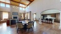 This elegant clubroom in Lubbock, Texas, is a generously sized gathering space designed with refined, upscale details throughout. The most striking architectural feature is the dramatic vaulted ceiling, finished with warm wood planking and clerestory windows that flood the interior with natural light. Rich hardwood flooring runs throughout, tying together the open, flowing layout.

The main living area centers around a stone fireplace with a classic mantel, complemented by a mounted flat-screen television above. Comfortable seating, including plush sofas and accent chairs, is arranged around a substantial coffee table, creating an inviting lounge atmosphere. Tall, floor-to-ceiling draperies frame the large windows, which offer views of the surrounding area.

A large round dining table with upholstered chairs occupies the central gathering zone, ideal for group meetings or social events. Elegant columned archways define the transition into the full kitchen, which features dark wood cabinetry, granite countertops, stainless steel appliances, a decorative patterned tile backsplash, and a bar-height island with seating. A chandelier with an artistic branch-like design adds a distinctive touch overhead.

The space is further enhanced by wall sconces, layered lighting, and thoughtful furnishings that collectively create a warm, sophisticated ambiance suited for community gatherings and events.