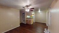 This single-story unit in Huntsville, Texas offers a functional and comfortable layout across its one-bedroom, one-bathroom floor plan. The open living area features dark hardwood-style flooring and a textured ceiling with a ceiling fan, creating a practical yet inviting atmosphere. Built-in shelving flanks the kitchen entrance, providing convenient display and storage space.

The kitchen is designed for efficiency, featuring warm oak cabinetry, light countertops, a double-basin sink, and a full suite of white appliances including a refrigerator, dishwasher, microwave, and range. A peninsula with a breakfast bar and two bar stools connects the kitchen to the living area, making it ideal for casual dining.

A second living space or dining area benefits from direct outdoor access through a glass-paneled door, accompanied by a large window with blinds that allow natural light to filter in.

The bedroom is carpeted for comfort, includes a ceiling fan, and features a window overlooking greenery. A hallway leads to the bathroom, which is equipped with a wood-toned vanity, a large mirror, and a well-lit vanity fixture, completing this practical and efficiently designed residence.