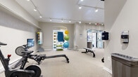 This fitness center at 261 College St in New Haven, CT, offers a thoughtfully designed single-floor workout space that balances functionality with a clean, modern aesthetic. The interior features neutral gray walls, plush carpeted flooring, and track lighting mounted along white ceilings, creating a bright and welcoming environment throughout.

The space is organized into distinct workout zones. A cardio area houses Echelon rowing machines, stationary bikes with interactive screens, treadmills, and an elliptical, all equipped with built-in displays for connected workouts. An ornate architectural frieze detail is visible near the ceiling in this section, hinting at the building's historic character.

A strength training area features a multi-station cable machine, a full dumbbell rack with a range of weights, and an adjustable workout bench. Wall-mounted mirrors enhance the sense of space and allow for form checks during training.

A functional training alcove, framed by an arched doorway, stores a colorful collection of stability balls, resistance tools, and exercise accessories. A small built-in desk near the glass entry door provides a convenient check-in or administrative area. Street-facing windows and a glass entrance allow natural light to filter in, connecting the space to the surrounding College Street streetscape.