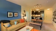 This single-floor residence showcases a thoughtfully designed open-concept layout that seamlessly integrates the living, dining, and kitchen areas into one cohesive and inviting space. The living area draws attention with a bold blue accent wall, warm neutral tones, and plush seating accented by colorful throw pillows, creating a relaxed yet stylish atmosphere. Framed artwork adorns the walls, adding a curated, personal touch. Adjacent to the living area, the dining space features a rustic wooden dining table with bench seating, making it both functional and visually appealing. Track lighting overhead provides ample illumination, enhancing the warmth of the space. The kitchen is well-appointed with sleek dark cabinetry, a stainless steel refrigerator, built-in microwave, and a peninsula with pendant lighting — ideal for both cooking and casual entertaining. Neutral-toned flooring flows throughout, tying the living and dining areas together harmoniously. The overall ambiance is modern and welcoming, with a careful balance of bold design choices and neutral backdrops that gives the space a vibrant yet comfortable character.
