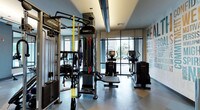 This fitness center offers a well-appointed, single-floor layout designed to accommodate a comprehensive range of workout routines. The centerpiece of the space is a Life Fitness Synrgy360 functional training system, featuring an array of resistance bands, TRX suspension straps, cable pulleys, and a rope attachment, making it suitable for full-body functional training. Accompanying this system is a substantial weight stack and adjustable bench, providing strength training versatility.

Cardio equipment, including elliptical machines and upright bikes, is positioned along the perimeter near floor-to-ceiling glass windows and sliding doors, allowing ample natural light to flood the space and offering views of the surrounding exterior. The open layout ensures ease of movement between stations.

The interior is finished with a light-toned, durable gym flooring, smooth gray and blue-accented walls, and recessed lighting complemented by a tray ceiling detail. A motivational typographic mural adorns one wall, reinforcing an energetic atmosphere. Full-length mirrors along one side enhance the sense of space while serving a functional purpose for form monitoring during exercise.