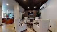 This thoughtfully designed single-floor amenity space blends contemporary style with warm, inviting materials throughout. The grand lobby entry sets the tone immediately, featuring polished concrete floors that reflect the ambient glow of recessed lighting, a striking slatted wood ceiling, and a grid of architectural wall sconces that create a dramatic yet welcoming first impression. Glass entry doors allow natural light to filter in, while elevators and a seating bench offer functional comfort.

Beyond the lobby, a refined lounge area serves as a central gathering space. A bold slatted dark wood accent wall anchors the room, framing a wall-mounted television above a sleek linear electric fireplace. Plush, nail-head-trimmed wingback chairs and a round coffee table complete the seating arrangement, exuding a club-like warmth and sophistication. Lush indoor greenery adds a natural, refreshing contrast to the rich wood tones.

A café-style refreshment bar features warm wood cabinetry topped with granite countertops, a professional espresso machine, and a vibrant deep-blue hexagonal tile backsplash accented by open industrial shelving adorned with plants and accessories.

The dining area showcases rich oak panel wainscoting, upholstered booth seating with leather strap details, and white marble-top tables. Framed vintage black-and-white sports photography lines the walls above, lending a timeless, curated character to the space. A separate conference area with a large communal table rounds out this well-appointed environment.