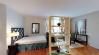 This single-floor studio is thoughtfully laid out to maximize functionality and comfort within an open-plan design. The main living area features warm parquet hardwood flooring throughout and is cleverly divided using freestanding shelving units that create distinct sleeping, living, and dining zones without sacrificing the sense of openness. Large windows allow generous natural light to fill the space, complemented by sheer curtains that soften the ambiance. The sleeping area is separated from the living space by a decorative room divider, providing a degree of privacy while maintaining an airy feel. A cozy seating area is anchored by a plush area rug, with wall art and accent furnishings adding character to the neutral-toned walls. A compact dining nook with bar-height seating sits adjacent to the kitchen area. The kitchen is efficiently designed with warm wood cabinetry extending to the ceiling for ample storage, a gas range, a full-size refrigerator, and a sink — all set atop the same parquet flooring. The bathroom is clean and functional, featuring classic white tile, a combination shower and bathtub, a pedestal sink, and a patterned floor tile that adds a subtle decorative touch. Overall, the space offers a cohesive, practical, and inviting environment.