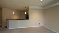 This single-floor space showcases a thoughtfully designed open-concept layout that connects the living area and kitchen in a seamless, functional flow. The living area features soft, neutral-toned carpet underfoot and warm beige walls that create a calm and welcoming ambiance. A raised kitchen bar with a dark countertop serves as both a practical workspace and a natural divider between the two areas, offering bar-style seating potential. The kitchen is equipped with dark cabinetry and a black refrigerator, giving it a sleek, contemporary feel. Track lighting in the kitchen, along with elegant pendant lights suspended above the bar, provide layered illumination that enhances the overall aesthetic. A recessed ceiling detail in the living area adds architectural interest and subtle dimension to the space. The open plan design maximizes the sense of space, making the interior feel airy and connected. Neutral tones throughout — from the carpeting to the walls — offer a versatile backdrop that accommodates a wide range of furnishing styles. Overall, the space combines practical functionality with clean, modern design elements to deliver a comfortable and visually cohesive environment.