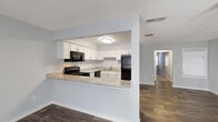 This single-story residence in East Lake, Florida, presents a clean, neutral interior designed for functional living. The open-concept layout connects the living and dining areas seamlessly, with dark hardwood-style flooring running throughout the main living spaces and a textured ceiling overhead. A distinctive white vertical-slat divider adds architectural interest, partially defining the living area while maintaining an open, airy feel. Large sliding glass doors and vertical blinds allow natural light to filter generously into the living space.

The kitchen is positioned as a pass-through, anchored by a granite-style countertop breakfast bar that opens toward the dining area. White shaker-style cabinetry provides ample storage, complemented by black appliances including a range, microwave, refrigerator, and dishwasher. The layout is efficient and well-organized.

The bedroom features plush carpeting and is generously equipped with dual sliding-door closets offering substantial storage. An en-suite bathroom includes a tub-shower combination, a white vanity with a granite-style countertop, and wood-style flooring. A second full bathroom with similar finishes serves the remaining areas of the home. Throughout, the soft gray walls and bright white trim create a consistent, contemporary aesthetic.