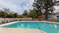This single-story community amenity space centers around a generously sized outdoor swimming pool with clearly marked depth zones, offering a refreshing recreational retreat. The pool deck is finished in light-colored concrete and lined with multiple sling-style lounge chairs, providing ample space for sunbathing and relaxation. A vibrant orange umbrella shades a picnic-style table and bench seating area, ideal for casual outdoor dining or socializing. The pool is enclosed by a neutral-toned privacy fence, lending a sense of seclusion while tall, mature pine and hardwood trees frame the backdrop, creating a natural, park-like atmosphere. Thoughtfully maintained landscaping, including ornamental grasses, trimmed shrubs, potted plants, and mulched garden beds, lines the perimeter of the deck, adding color and texture throughout. Decorative lamp posts punctuate the space, providing ambient lighting for evening use. A covered entry corridor with a blue-painted tongue-and-groove ceiling leads to the pool area through glass sliding doors, while a small alcove nearby houses a Coca-Cola vending machine for added convenience. Overall, the space strikes a balance between functional amenity and pleasant outdoor living.