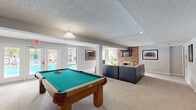 This single-floor game room is a versatile and well-designed entertainment space that seamlessly blends recreational and lounge areas into one cohesive layout. The room is divided into two functional zones: a billiards area centered around a solid oak pool table with a teal felt surface, illuminated by a classic pendant light overhead, and a comfortable lounge area outfitted with a tufted gray sectional sofa and accent chairs arranged around a wall-mounted flat-screen TV. A striking brick fireplace serves as the focal point of the lounge zone, adding warmth and a rustic architectural contrast to the otherwise contemporary, light gray palette throughout. The space features plush wall-to-wall carpeting, a textured ceiling, and recessed lighting that creates a bright and inviting atmosphere. A wall-mounted cue rack keeps billiards equipment neatly organized, while framed artwork adorns the walls throughout. Large windows and glass double doors flood the interior with natural light and offer direct views of an outdoor pool area, creating a strong indoor-outdoor connection. A set of double interior doors provides additional storage or access, rounding out the practical design of this well-equipped game room.