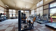 This spacious fitness center is thoughtfully designed to support a wide range of workout routines in a bright, welcoming environment. The room features striking vaulted ceilings that soar overhead, enhancing the sense of openness and airiness throughout the space. Large windows with adjustable roller shades line the walls, allowing generous amounts of natural light to filter in while offering views of the surrounding outdoor greenery.

The facility is comprehensively equipped for both cardio and strength training. A row of treadmills and elliptical machines lines one side of the room, providing ample options for cardiovascular exercise. On the strength training side, multiple cable and resistance machines with tan leather upholstery are positioned throughout the floor, complemented by a dedicated free weights area stocked with a full rack of dumbbells in varying sizes. A functional training corner houses an impressive collection of medicine balls, slam balls, kettlebells, and battle ropes, catering to dynamic and circuit-style workouts.

The interior design features warm taupe walls accented by dark wood trim details and framing elements, lending the space a refined, lodge-inspired aesthetic. Patterned carpet tiles in neutral tones cover the floor, adding texture and visual interest. Ceiling fans and pendant lighting fixtures provide additional illumination and comfort. A wall-mounted television is also present, rounding out this well-appointed fitness facility.