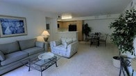 This single-floor space offers a thoughtfully designed open-concept layout that seamlessly connects the living, dining, and kitchen areas into one cohesive and functional environment. The living area is anchored by a plush sofa and a generously sized armchair, both upholstered in soft, neutral tones, arranged around a glass-topped coffee table with an ornate metal base. Warm ambient lighting from a classic table lamp complements the recessed and track lighting overhead, creating a comfortable and inviting atmosphere throughout. Soft wall-to-wall carpeting in a light neutral tone extends across the main living space, adding warmth underfoot. The dining area sits adjacent to the living room and features a round glass table paired with elegant wrought-iron chairs, ideal for intimate gatherings. A lush potted plant nearby adds a natural, organic touch to the space. The kitchen is set back slightly from the main living area, defined by light wood cabinetry, a black refrigerator, and a raised breakfast bar that serves as both a functional workspace and a casual seating option. The clean, neutral color palette and efficient use of space make this a practical and comfortable single-floor residence.