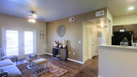 This single-floor residence offers a cohesive, open layout designed for comfortable everyday living. The main living area is anchored by rich, dark hardwood-style flooring that extends throughout the space, complementing the warm beige-toned walls. A ceiling fan with a light fixture provides both airflow and ambient lighting, while a window along one wall invites natural light into the room. Decorative wall art and a console table with curated accents add character to the living space, which also features a plush area rug and seating.

A wide opening leads from the living area into a hallway corridor, which connects to additional rooms, including what appears to be a bedroom with a louvered closet door. The hallway is well-lit and displays wall art, contributing to the home's cohesive aesthetic. A partial view of the kitchen reveals light-toned countertops, suggesting a functional and well-appointed cooking space. HVAC vents are positioned throughout the ceiling, ensuring consistent climate control. The overall ambiance is warm, neutral, and inviting, with a clean, move-in-ready feel throughout.