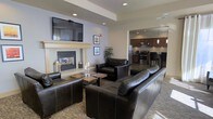This single-floor space offers a thoughtfully designed communal environment that blends comfort and functionality. The central lounge area is anchored by a classic fireplace with a crisp white mantel, above which a wall-mounted flat-screen TV is positioned for easy viewing. Plush dark leather sofas and armchairs are arranged around a rustic wooden coffee table, all set atop a patterned area rug, creating a warm and inviting atmosphere. Recessed lighting throughout the space provides consistent, well-distributed illumination, while neutral wall tones contribute to a clean, modern aesthetic. Framed artwork adds subtle character to the walls, enhancing the overall ambiance. Adjacent to the lounge, an open kitchen area features dark wood cabinetry, stainless steel appliances including a gas range and refrigerator, and pendant lighting that hangs above what appears to be a bar or counter seating area. The open-plan layout creates a seamless visual connection between the lounge and kitchen, making the space well-suited for both relaxed gatherings and casual dining. The overall design balances contemporary style with approachable comfort, making it a functional and welcoming shared environment.