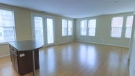This single-floor space is characterized by its open, airy layout and an abundance of natural light. The main living area showcases smooth hardwood-style flooring in a warm honey tone, which reflects light throughout the room and contributes to the overall sense of spaciousness. Soft, neutral-toned walls in a light sage-green hue complement the clean white ceiling and trim, creating a calm and inviting atmosphere.

A prominent set of French doors along one wall provides both a visual focal point and direct access to what appears to be an outdoor area or balcony, offering views of the surrounding building exterior. Flanking the French doors and continuing along the adjacent wall are several large windows fitted with white blinds, ensuring consistent natural light from multiple angles throughout the day.

The open floor plan allows for flexible furniture arrangement, making the living area highly versatile. Clean white baseboards and subtle architectural details, including visible air vents and standard outlets, reflect a practical, modern design sensibility. A partial view of a dark-toned table edge suggests a dining or workspace area nearby, hinting at a functional layout suited to everyday living.