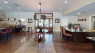 This single-story clubhouse is designed to serve as a welcoming community gathering space, thoughtfully laid out to accommodate a variety of activities and social occasions. Rich hardwood flooring flows throughout the open-concept interior, lending warmth and continuity across all areas.

The main lounge area is anchored by a striking stacked-stone fireplace with a wall-mounted television, creating an inviting focal point. A sweeping curved wall of large windows frames direct views of the outdoor pool and patio area, flooding the space with abundant natural light. Comfortable seating arrangements and decorative greenery contribute to the relaxed yet polished ambiance.

The entry foyer features a stylish round wood accent table beneath a modern drum pendant light fixture, with wood-framed double doors providing an elegant arrival experience. A dining area with upholstered seating is positioned nearby, ideal for meetings or casual gatherings.

The clubhouse kitchen is fully equipped with dark espresso cabinetry, granite countertops, a stainless steel double sink, a full-size French door refrigerator, and a dishwasher — making it well-suited for catered events or resident use. Adjacent to the kitchen, a glass-paned door leads to a dedicated fitness room stocked with free weights and exercise equipment, offering residents convenient on-site wellness facilities.