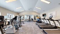 Located at 4007 Raynor Pkwy in Bellevue, Nebraska, this fitness center offers a generously sized, single-floor exercise room designed to support a comprehensive workout experience. The space features striking vaulted ceilings that lend an open, airy feel, complemented by recessed panel lighting and a ceiling fan that keep the environment bright and comfortable. Soft sage green and neutral-toned walls contribute to a calm, focused atmosphere throughout.

The exercise room is well-appointed with a wide array of cardio equipment, including multiple TRUE-brand treadmills, elliptical machines, and stationary bikes, providing ample options for aerobic training. A dedicated strength training area features a multi-function cable machine, a dumbbell rack with a full weight selection, and an adjustable workout bench. Stability balls are also available for functional fitness exercises.

Large windows with blinds allow natural light to flow into the space while maintaining privacy, and a floor-to-ceiling mirror along one wall visually expands the room and aids users in monitoring their form. Wall-mounted flat-screen televisions are positioned for convenient viewing during workouts. The durable, patterned carpet tiles underfoot provide both comfort and traction. Additional rooms accessible from the main floor suggest supplementary amenities nearby, rounding out this fully functional fitness facility.