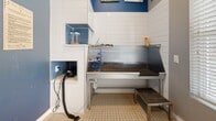 This purpose-built dog wash station is a compact, well-appointed space designed specifically for pet grooming and bathing. The room is finished with clean white subway tile along the grooming wall, complemented by bold blue accent walls that lend a lively, welcoming character to the space. The flooring features small-format mosaic tile, practical for wet conditions and easy maintenance.

The centerpiece of the room is a raised stainless steel grooming tub equipped with a coiled hose and spray attachment, providing efficient water access for bathing pets of various sizes. A step stool positioned at the tub offers convenient access. Built into the tiled surround are dedicated recessed compartments, including one housing a mounted blow dryer with hose, as well as shelving niches with blue penny-tile detailing for storing grooming supplies.

Natural light enters through a window fitted with white blinds, brightening the space alongside overhead lighting. A glass-panel exterior door provides additional light and direct outdoor access. The room is accessed through elegant double panel doors with brushed nickel hardware, adding a polished touch to the functional space. Charming dog-themed artwork displayed on the upper blue accent wall adds a warm, inviting finishing detail to this thoughtfully designed pet grooming area.