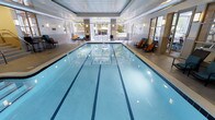 This premier indoor aquatic facility is designed to offer both functionality and comfort in a refined setting. At its centerpiece is a spacious, clearly marked multi-lane lap pool with crisp, blue water and underwater lighting that enhances visibility and ambiance. The pool floor features a prominently tiled club logo, adding a polished, branded touch to the space.

The interior is characterized by a dropped ceiling with recessed lighting and elegant wall sconces that provide warm, even illumination throughout. Large windows along the far wall flood the space with natural light and offer views of lush greenery outside, creating a seamless connection between the indoor environment and the surrounding landscape.

Along the pool deck, a selection of lounge chairs and seating areas are thoughtfully arranged to provide comfortable spaces for relaxation before or after a swim. The neutral color palette of the walls and flooring complements the clean, contemporary architectural aesthetic of the space.

The facility maintains a calm and resort-like atmosphere, balancing practical amenities with an upscale design sensibility. Whether used for dedicated lap swimming or leisurely aquatic activities, this indoor pool facility delivers a high-quality experience in a well-appointed environment.