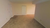 This single-floor space presents a straightforward and functional layout with an open, uncluttered interior. The floors throughout are covered in wall-to-wall carpeting in a warm beige tone, providing a soft underfoot feel across the main living area. The walls are finished in a neutral cream shade, lending a clean and understated backdrop that would accommodate a variety of décor styles. A classic six-panel white door serves as a focal point along one wall, offering access to an adjoining room or closet space. The ceiling features a drop-tile grid design, which is practical and commonly found in ground-level or below-grade spaces. The layout opens into an adjoining area around a partial wall partition, suggesting a flexible floor plan that could accommodate multiple functional zones. Baseboard trim lines the perimeter of the room, adding a subtle finishing detail. The overall ambiance is minimal and neutral, making it a blank canvas well-suited for customization. The space benefits from its open proportions, offering room to configure furnishings and functional areas according to individual needs.
