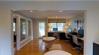 This single-floor space offers a thoughtfully designed interior that balances functionality with classic charm. The main area serves as a dedicated workspace, featuring a built-in desk running along the back wall that accommodates dual computer workstations, each equipped with monitors and complemented by elegant task lighting. Dark cabinetry beneath the desk provides ample storage while adding a refined contrast to the lighter walls and trim. Recessed ceiling lighting is evenly distributed throughout, ensuring consistent illumination across the entire space. Crown molding along the ceiling adds a touch of traditional architectural character. Natural light flows generously through multiple windows dressed with tailored Roman shades, as well as through a glass-paneled exterior door and large sidelight windows featuring decorative leaded glass detailing — a notable architectural element that lends warmth and visual interest. The warm-toned hardwood flooring extends throughout, adding to the inviting ambiance. A round table with modern chairs in the center of the room provides a comfortable area for informal meetings or collaborative work. The exterior views through the windows reveal a lush, tree-lined outdoor setting, further enhancing the pleasant atmosphere of this well-lit, professionally arranged space.