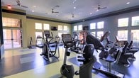 This fitness center offers a spacious, single-floor layout filled with a comprehensive selection of cardio equipment, including treadmills, elliptical trainers, stationary bikes, and stair climbers. The room is bright and welcoming, with large windows lining the walls on multiple sides, allowing generous natural light to flow throughout the space. The flooring features a two-tone design, combining light and dark sections that help define workout zones while adding visual interest. Ceiling fans are evenly distributed across the room, providing consistent airflow to keep the environment comfortable during workouts. Recessed lighting supplements the natural light, ensuring the space remains well-lit at any time of day. Mounted flat-screen televisions are positioned at convenient viewing angles, allowing users to stay entertained while exercising. The walls feature a calming two-tone color scheme, with a soft yellow-green on the lower portion and a muted teal-gray above, contributing to an energizing yet relaxed atmosphere. The layout is thoughtfully arranged to allow ample room between machines, making navigation easy and workouts comfortable. Overall, the space is clean, functional, and designed to support a productive fitness experience.