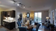 This single-floor space showcases a thoughtfully designed open-concept layout that connects the dining and living areas in a cohesive and elegant arrangement. The dining area is anchored by a rich, dark wood round table surrounded by tufted navy blue upholstered chairs, creating a sophisticated focal point. Above, a track lighting system with adjustable spotlights provides targeted, modern illumination throughout the space.

The living area flows naturally from the dining space, featuring a tufted sofa, a patterned area rug, and decorative accessories that add layers of texture and warmth. A round convex mirror serves as a striking wall accent, flanked by ornate globe-style floor lamps that cast a warm, ambient glow. A glass sliding door bathes the space in natural light and provides direct access to a private balcony with outdoor views, enhancing the sense of openness.

Dark hardwood-style flooring runs throughout, complementing the deep-toned cabinetry visible in the adjacent kitchen area. A glimpse into a secondary room reveals additional furnishings and décor, suggesting a well-appointed and cohesive interior design scheme throughout the entire residence.