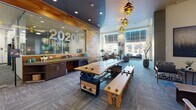 Located at 2020 Lawrence Street in Denver, CO, this thoughtfully designed amenity floor serves as the social and functional hub of the community. The welcoming lobby greets visitors with warm wood-planked accent walls, pendant lighting with an industrial flair, and a striking live-edge communal table — all combining to create an inviting, contemporary atmosphere.

The lounge area features tufted blue sofas anchored by a bold wood-slice area rug, centered around a wall-mounted TV set against rich horizontal wood paneling. Natural light pours in through floor-to-ceiling windows in the coworking zone, which offers multiple seating configurations, natural wood tables, and a deep blue accent wall with built-in media display — ideal for remote work or casual gatherings.

The full demonstration kitchen is a standout feature, outfitted with stainless steel appliances, a large range hood, mosaic tile backsplash, granite countertops, dark wood cabinetry, and a generous island with bar seating — perfect for resident events and entertaining. A dedicated package locker area provides added convenience, set beneath dramatic double-height ceilings adorned with abstract wall art. Throughout the space, polished concrete floors, curated greenery, and cohesive modern design elements create a vibrant and functional community environment.